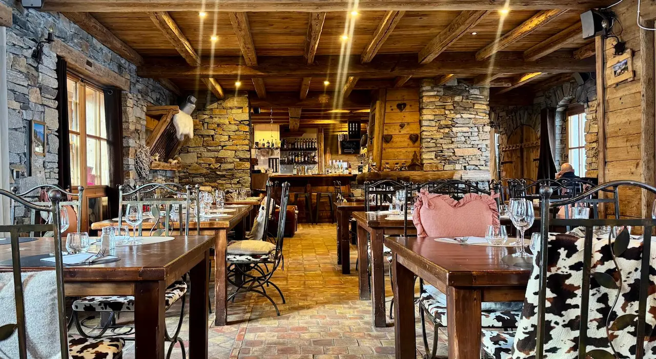 Rustic, dark, and warm chalet interior, with a large stone and wood fireplace in the center, and fur seating.