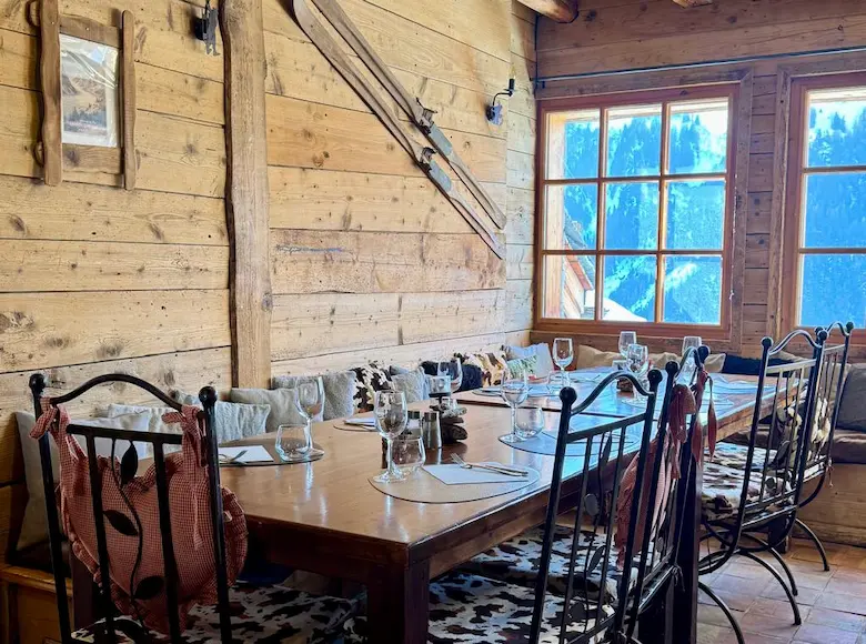 Fondue and charcuterie meal on a terrace table, with a village of chalets and a spectacular panorama of snowy alpine mountains under the sun in the background.