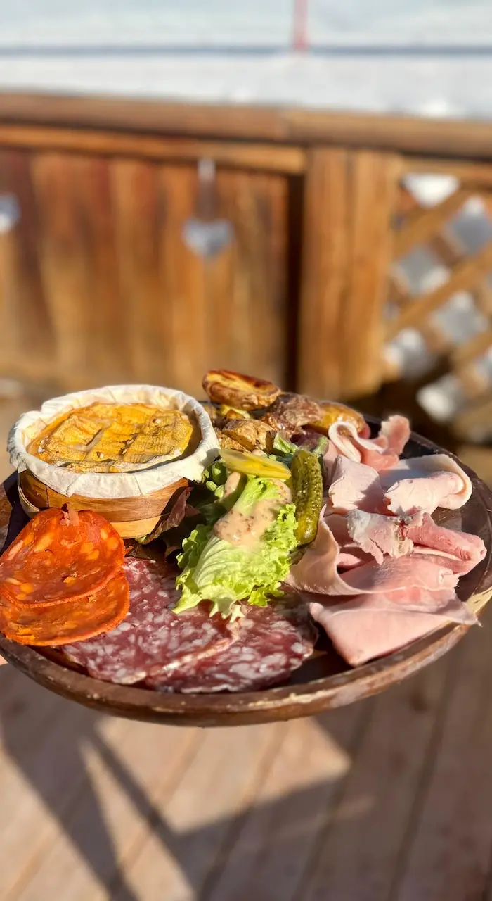 Wooden table set in a rustic chalet interior. The decor features wood and stone walls, wrought iron chairs with cowhide pattern cushions, and a beamed ceiling.