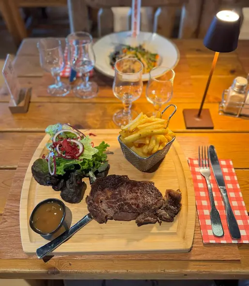 At the Le K2 restaurant, a smiling dark-haired barmaid, wearing a navy blue shirt and a brown leather apron, stands behind a rustic log wood bar counter. The back bar is filled with rows of liquor bottles and hanging glasses.