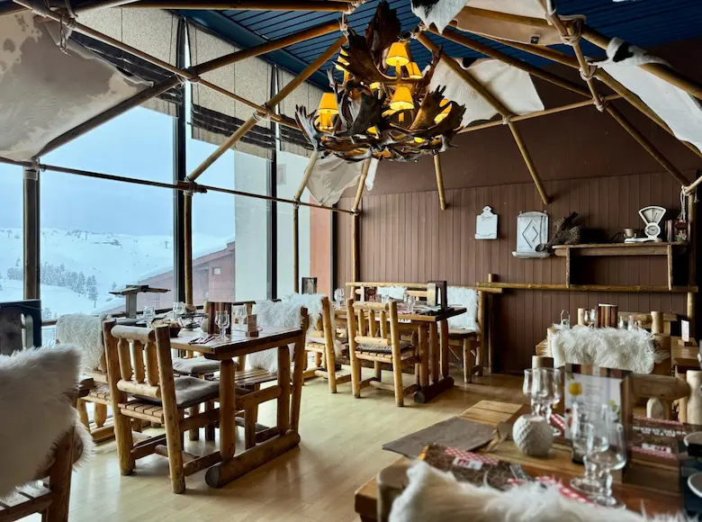 Wide view of the dining room of a restaurant called 'Le K2' with a mountain chalet decor. The tables and chairs are made of light wood and are adorned with white fur throws and cushions and a cushion decorated with a moose.