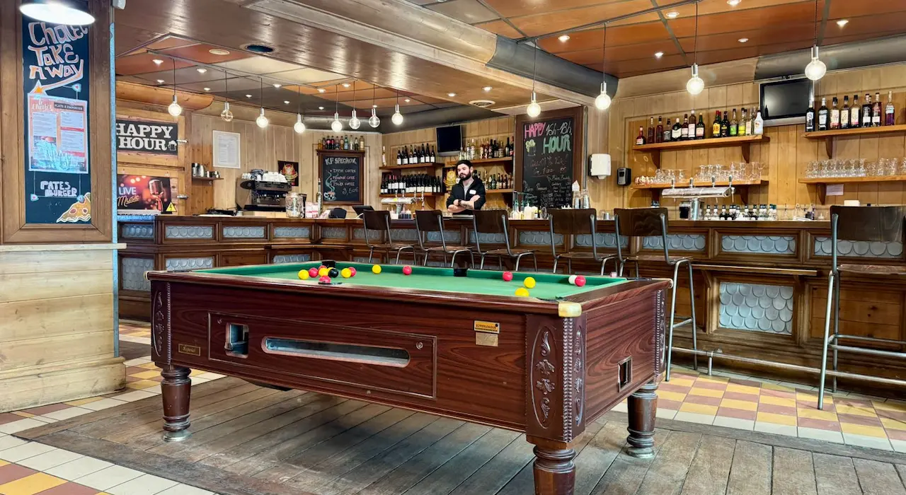 View of a wooden table set for raclette and charcuterie, with salad, potatoes, and three bottles of 'Savoie' red wine. The table is placed near a window overlooking a snowy landscape, in a wooden interior.