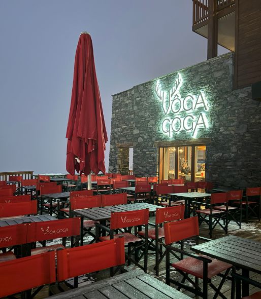 Snowy outdoor terrace of the VÔGA GOGA restaurant at dusk. The building is made of stone with a large bright sign. The terrace is furnished with lots of dark tables and folding red director chairs, with a large red umbrella closed in the center.