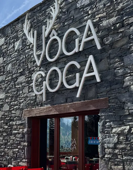 Exterior of a mountain chalet-style building, with a stone wall and a wooden terrace, displaying a large 'VOGA GOGA' sign with a deer antler logo. A red director's chair with the same logo is visible in the foreground, under a cloudy blue sky.