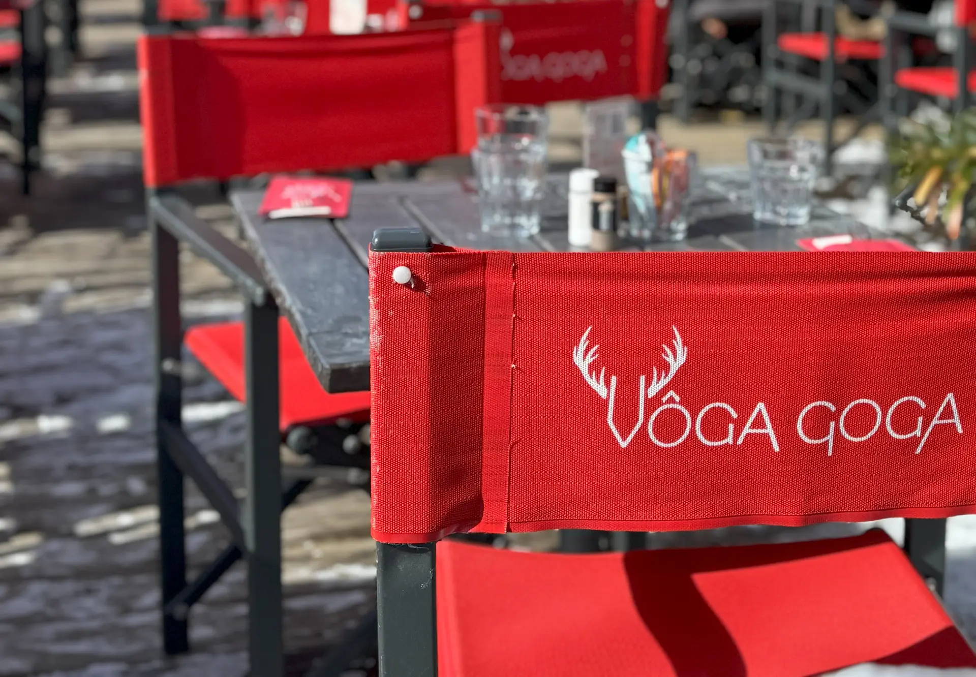 Gros plan sur l'enseigne 'VÔGA GOGA' en grandes lettres blanches modernes avec un logo de bois de cerf stylisé, fixée sur un mur extérieur de pierres grises. La porte d'entrée en bois et verre, affichant les mots 'PUB' et 'RESTAURANT', est visible à droite