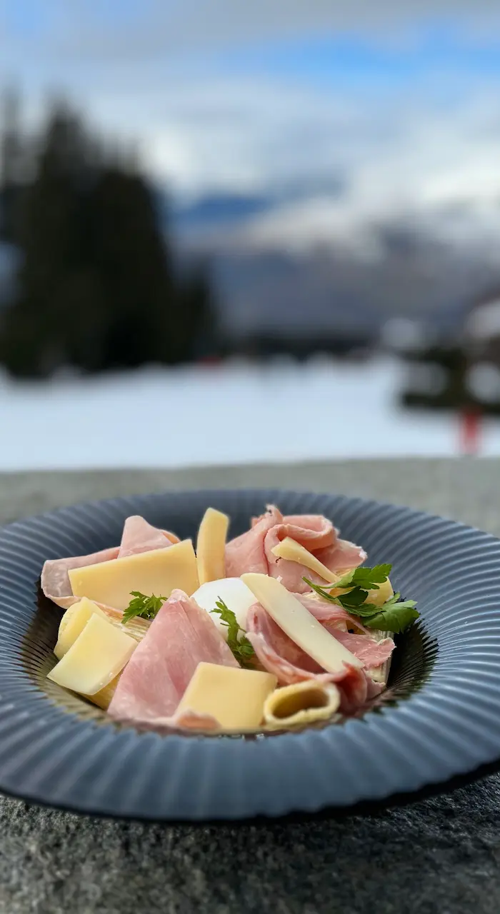 Un hamburger sur pain aux graines avec des frites présentées dans un cornet en papier journal et une salade d'accompagnement dans un bol blanc, posés sur une planche de bois sur la terrasse du restaurant, avec une vue panoramique des montagnes enneigées en arrière-plan.