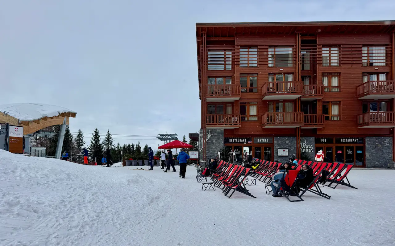 Gros plan sur l'enseigne 'VÔGA GOGA' en grandes lettres blanches modernes avec un logo de bois de cerf stylisé, fixée sur un mur extérieur de pierres grises. La porte d'entrée en bois et verre, affichant les mots 'PUB' et 'RESTAURANT', est visible à droite