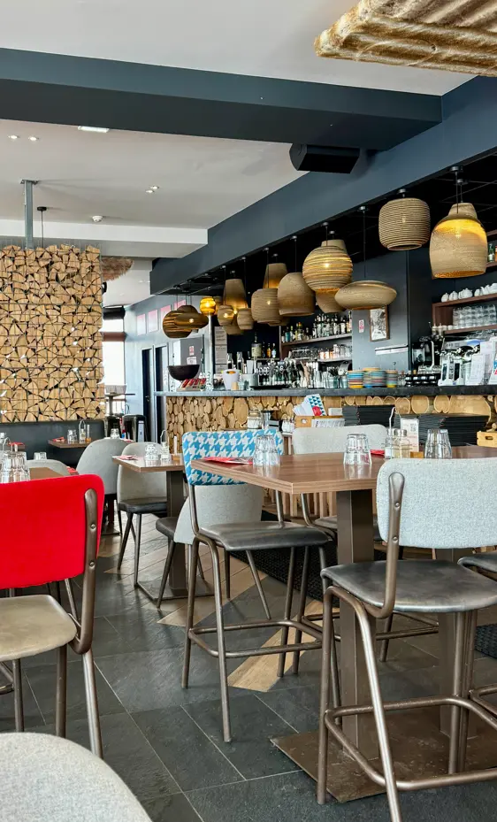 Un hamburger sur pain aux graines avec des frites présentées dans un cornet en papier journal et une salade d'accompagnement dans un bol blanc, posés sur une planche de bois sur la terrasse du restaurant, avec une vue panoramique des montagnes enneigées en arrière-plan.