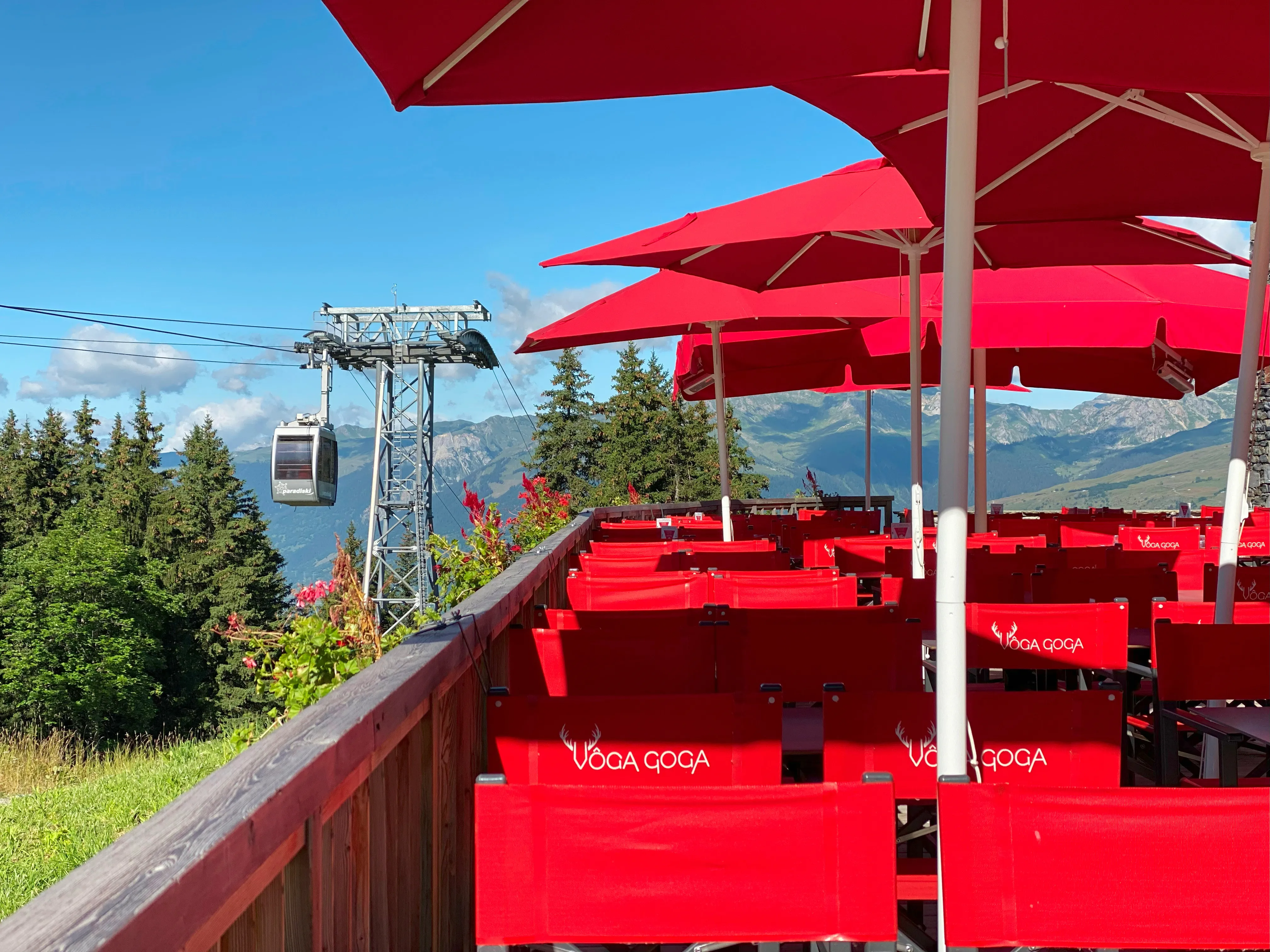 Grande table de terrasse rectangulaire du restaurant VÔGA GOGA, dressée pour plusieurs personnes avec des verres à eau et des serviettes de table rouges, encadrée de chaises de réalisateur rouges. En arrière-plan, vue panoramique sur les sommets des montagnes enneigées et un télésiège, sous un ciel bleu éclatant
