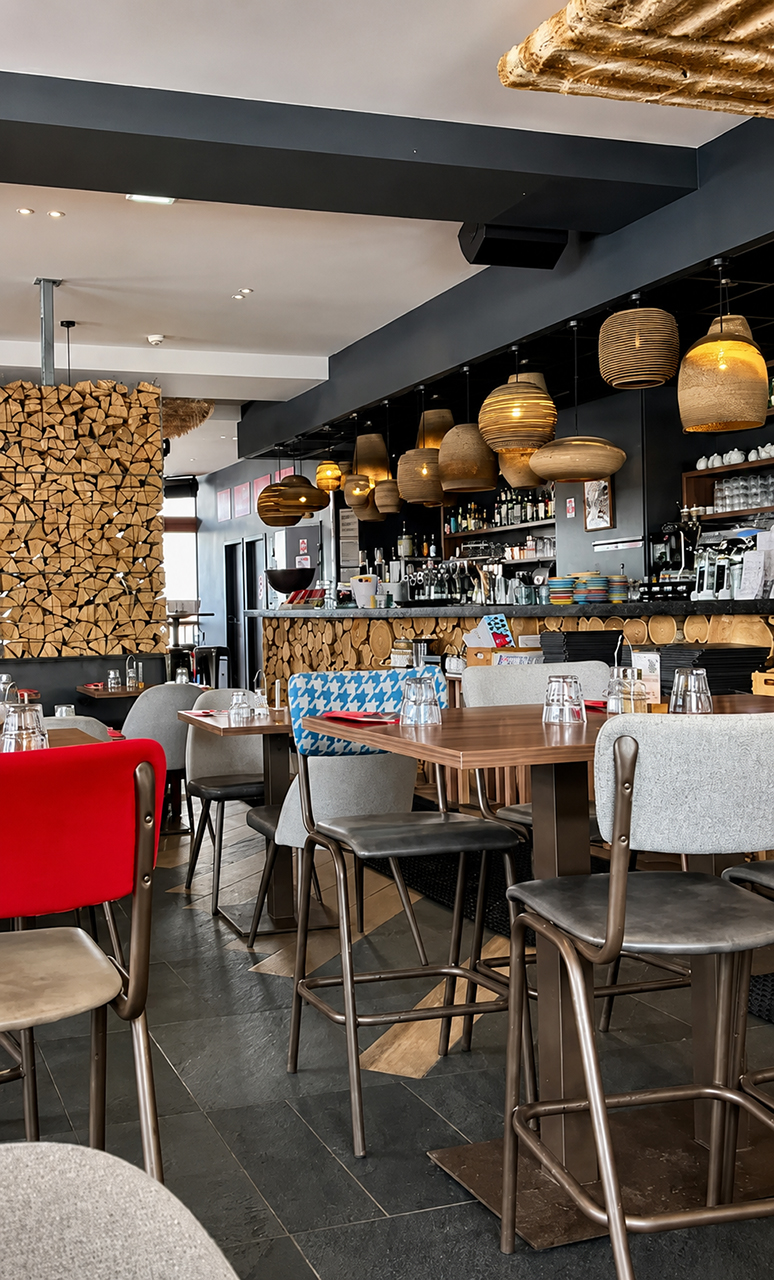 Un hamburger sur pain aux graines avec des frites présentées dans un cornet en papier journal et une salade d'accompagnement dans un bol blanc, posés sur une planche de bois sur la terrasse du restaurant, avec une vue panoramique des montagnes enneigées en arrière-plan.