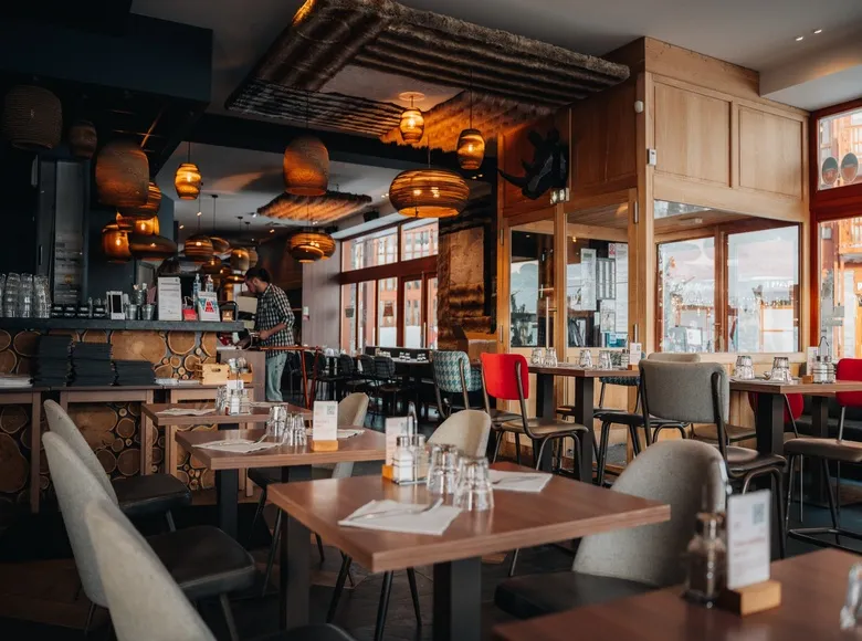 Gros plan sur l'enseigne 'VÔGA GOGA' en grandes lettres blanches modernes avec un logo de bois de cerf stylisé, fixée sur un mur extérieur de pierres grises. La porte d'entrée en bois et verre, affichant les mots 'PUB' et 'RESTAURANT', est visible à droite