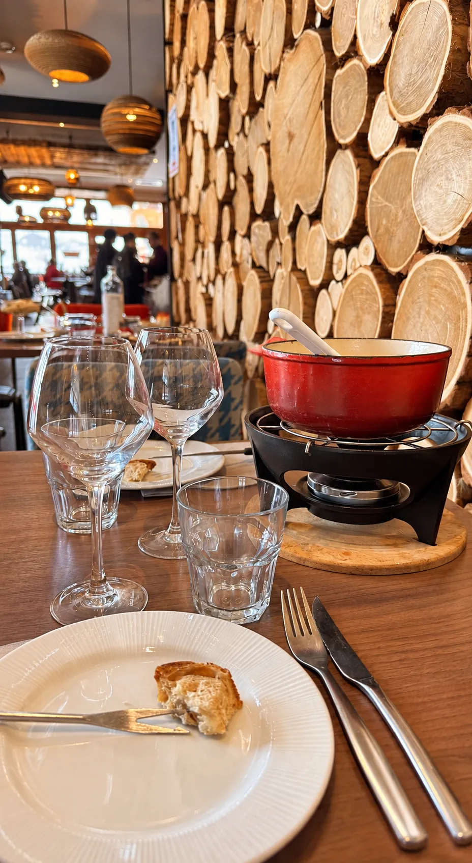 Un hamburger sur pain aux graines avec des frites présentées dans un cornet en papier journal et une salade d'accompagnement dans un bol blanc, posés sur une planche de bois sur la terrasse du restaurant, avec une vue panoramique des montagnes enneigées en arrière-plan.