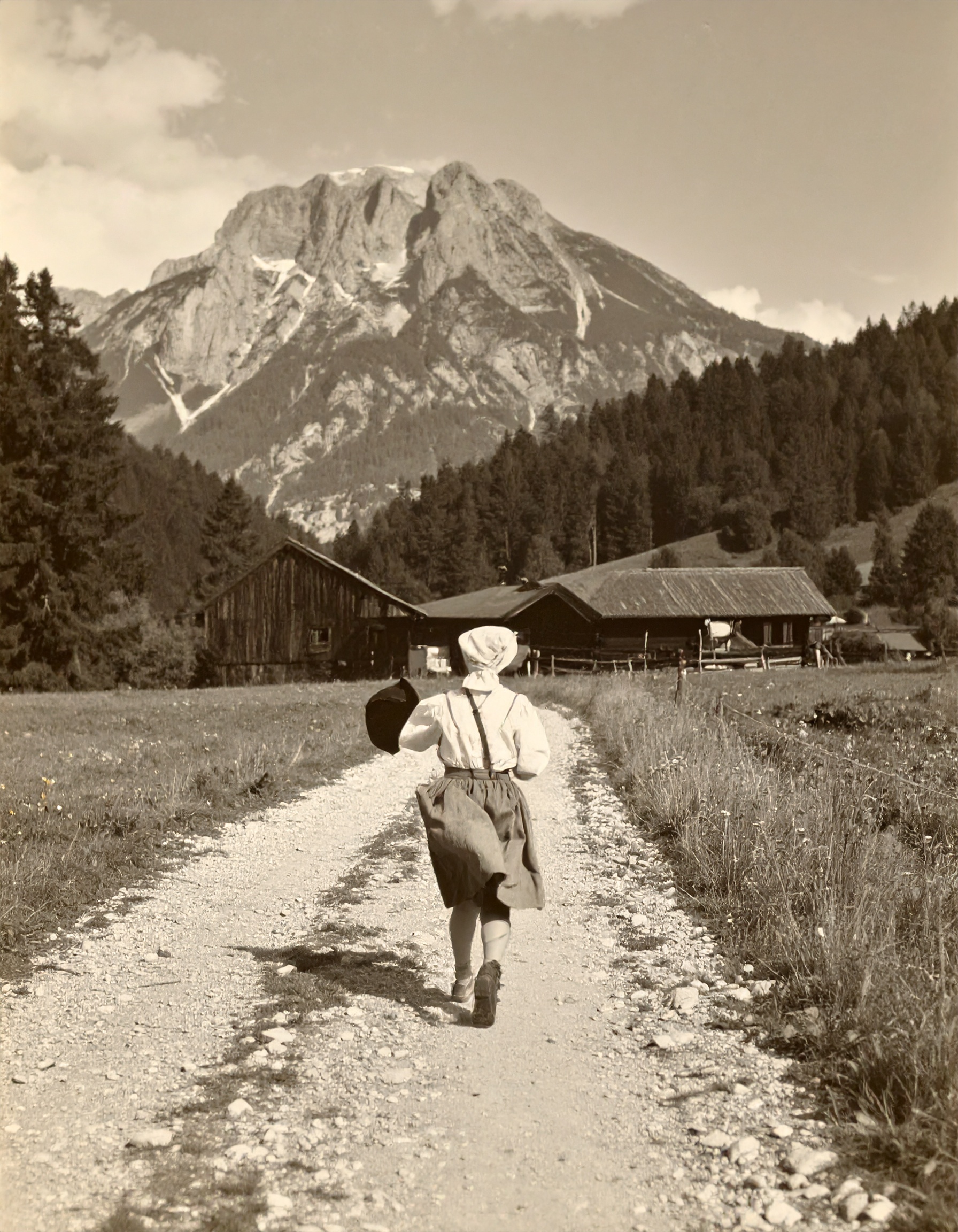Black-and-white photo of a girl in traditional attire walking on a gravel path towards wooden houses with a large mountain and forest in the background.