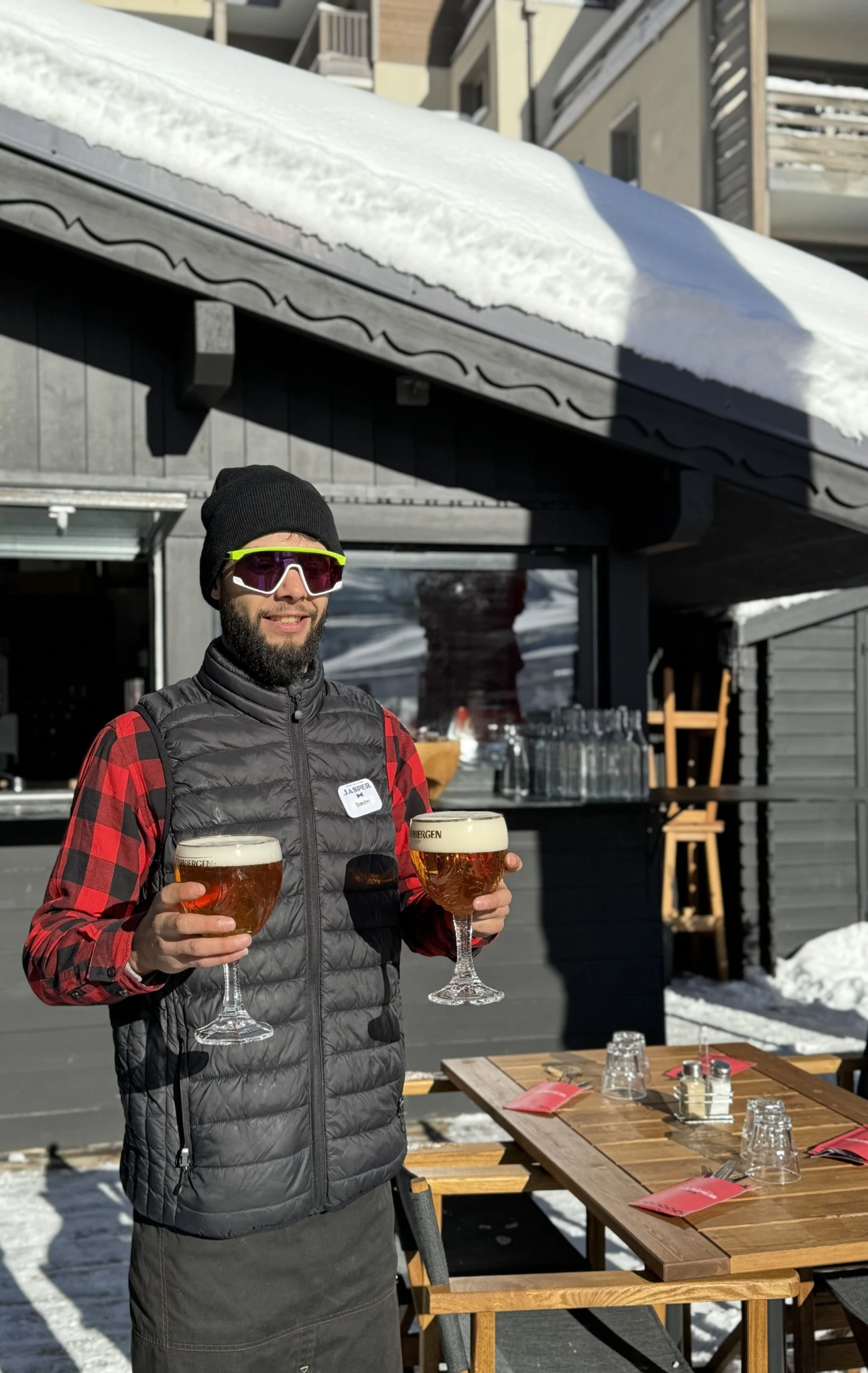 A man wearing a black vest and a cap, holding two glasses of beer, standing in front of the terrace of the restaurant Le Jasper.