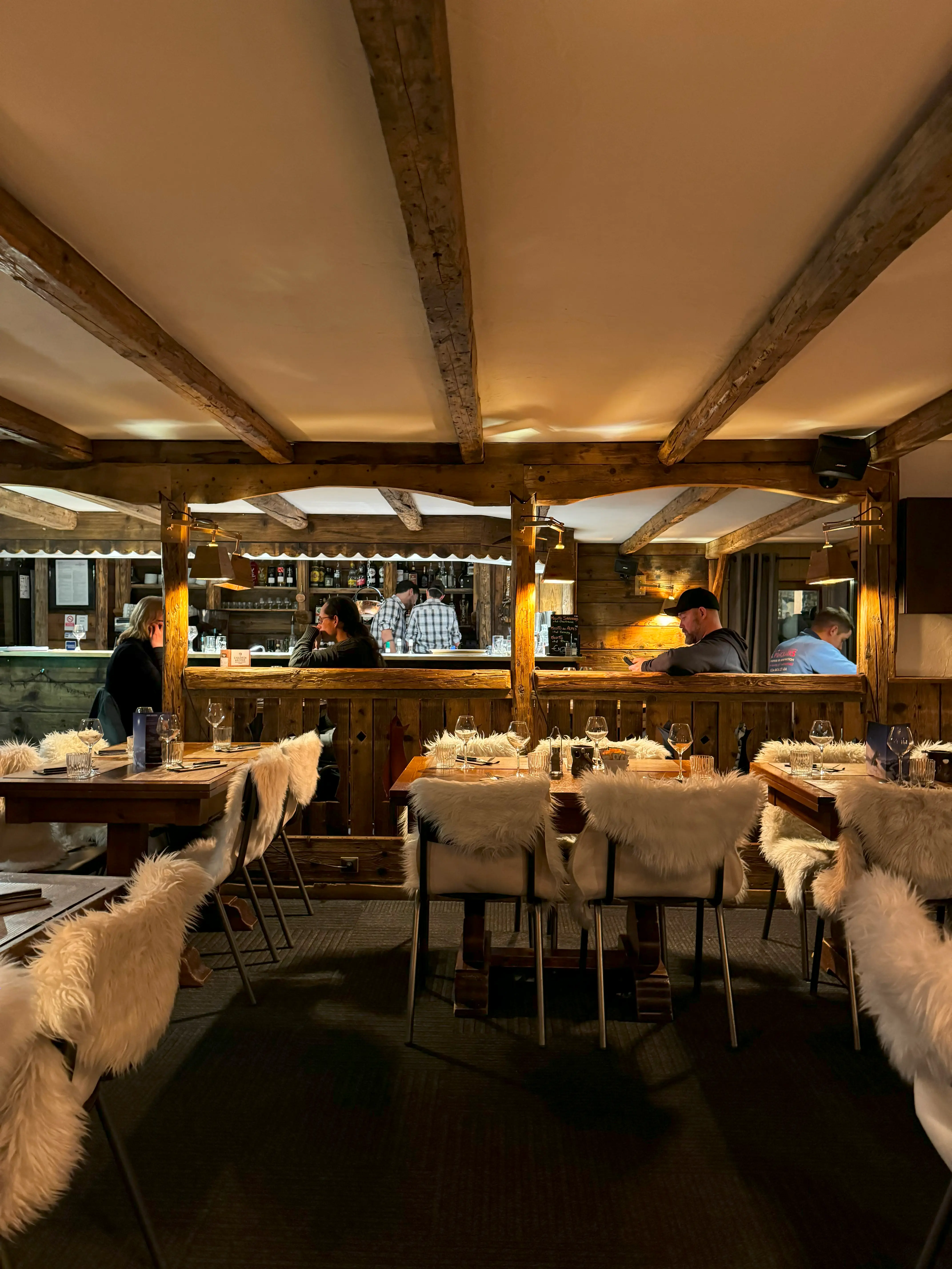 Cozy rustic restaurant interior with wooden beams, tables set with glassware, and chairs draped in white fur covers.