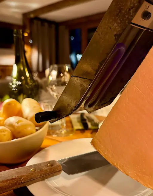 Intérieur chaleureux du restaurant Chaudron Magique, mettant en vedette un coin repas avec des murs et des poutres en bois brut, une longue table en bois, et des chaises de style industriel. L'ambiance est cosy et montagnarde, éclairée par des appliques murales et la lumière des fenêtres.