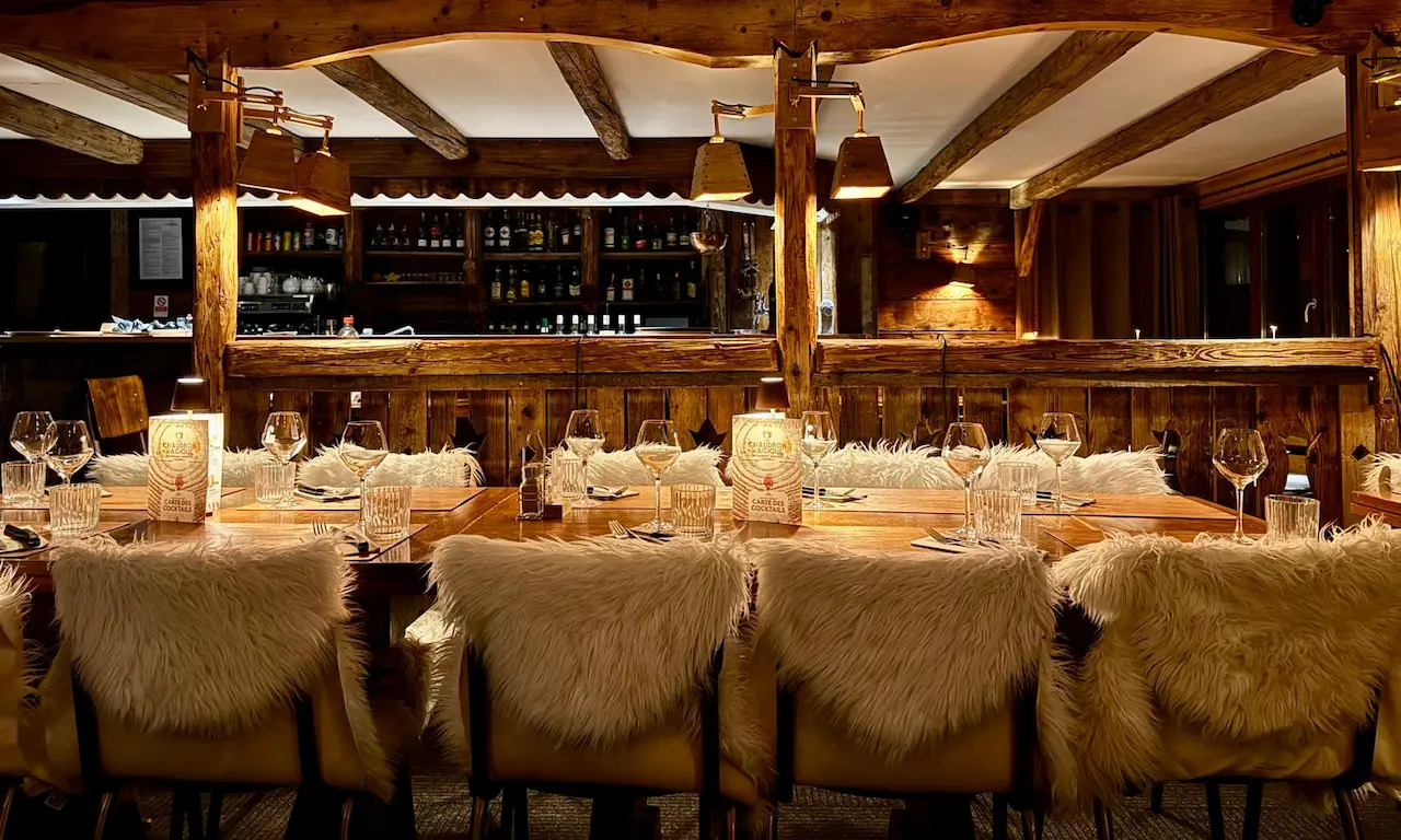 Salle de restaurant rustique et intime avec une longue table dressée, des murs et poutres en bois brut. Les chaises sont ornées de peaux ou de fourrures blanches pour une ambiance montagnarde et cosy. L'éclairage tamisé crée une atmosphère chaleureuse.