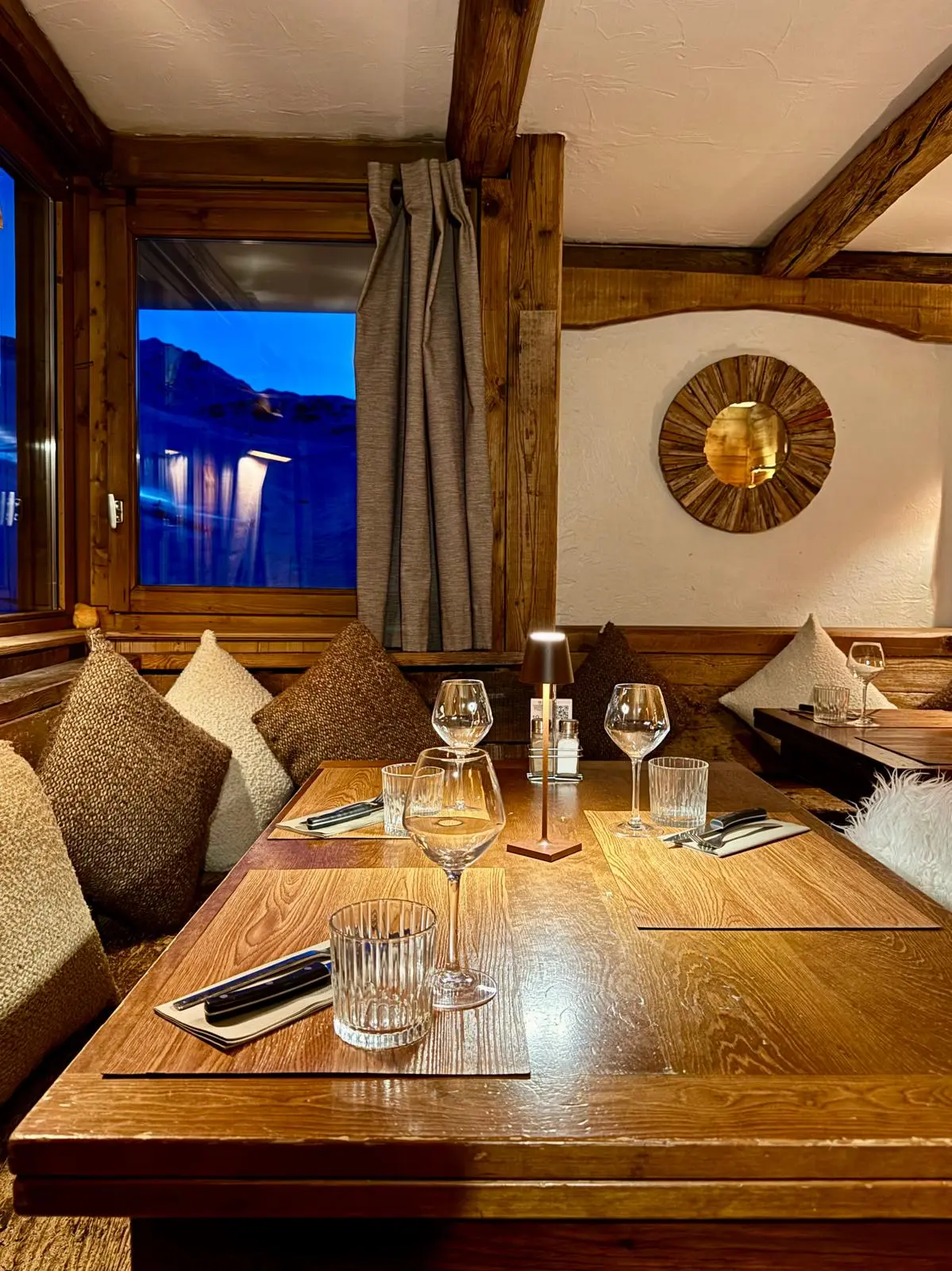 Table de restaurant dressée pour quatre personnes dans une ambiance chalet alpine. La salle est caractérisée par des murs en bois foncé et rustique, des chaises recouvertes de peaux de mouton blanches, et un éclairage suspendu tamisé. Des reflets d'une vitre ou d'un miroir encadré en bois sont visibles à l'arrière-plan.