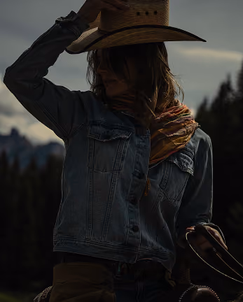 Person wearing a cowboy hat, denim jacket, and colorful scarf holding horse reins in an outdoor setting.