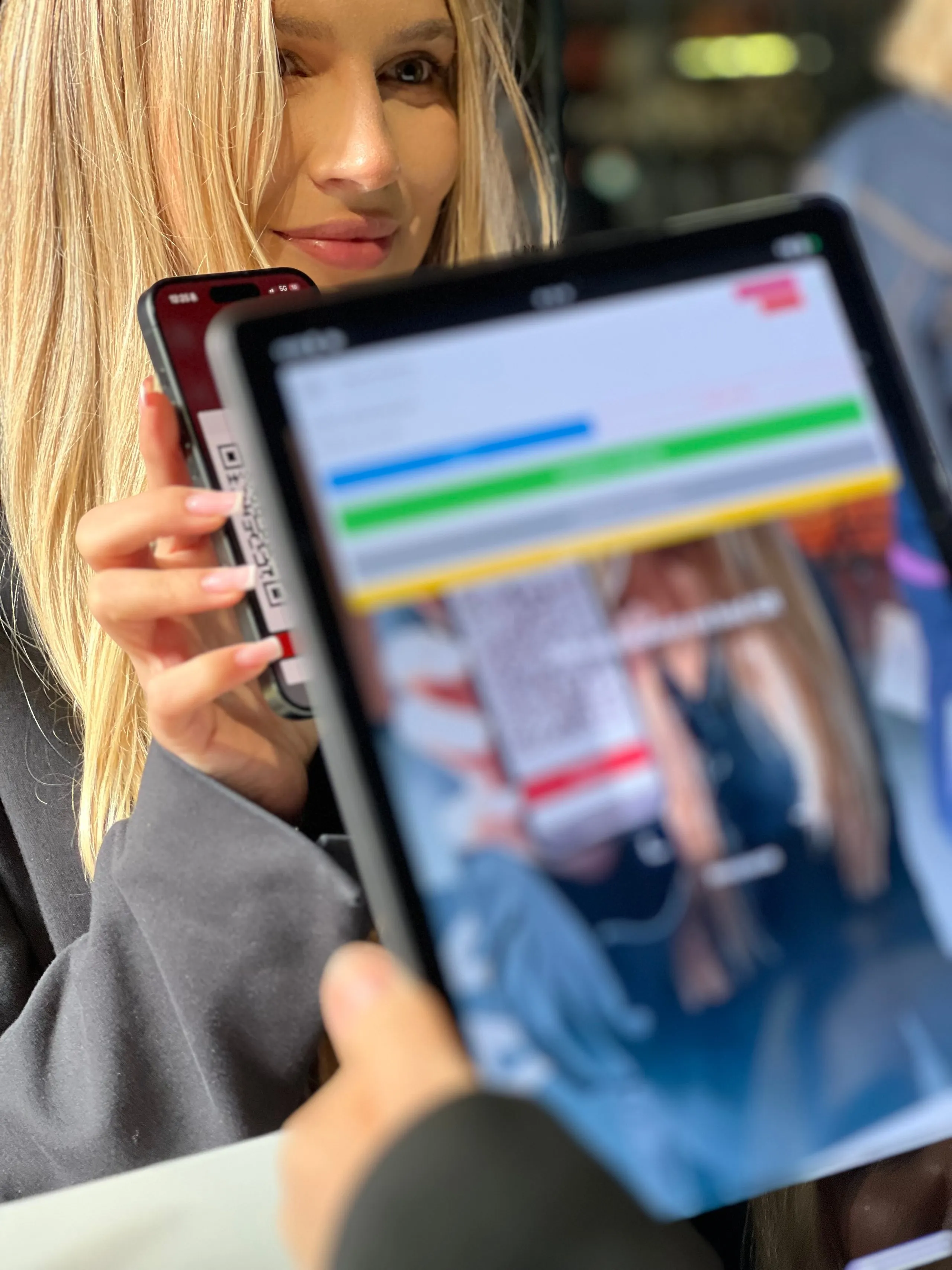 Młoda kobieta pokazuje ekran smartfona z kodem QR, podczas gdy ktoś inny skanuje go za pomocą tabletu.