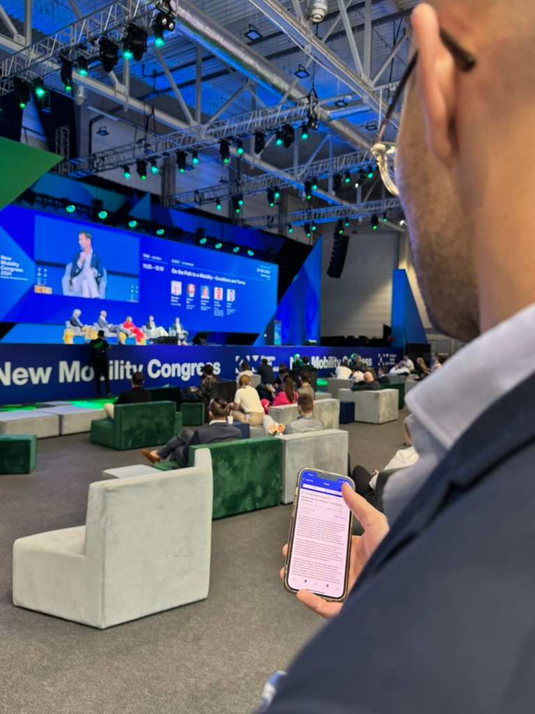 Mężczyzna w okularach trzyma smartfon i obserwuje panel dyskusyjny na dużej scenie podczas Kongresu Nowej Mobilności.
