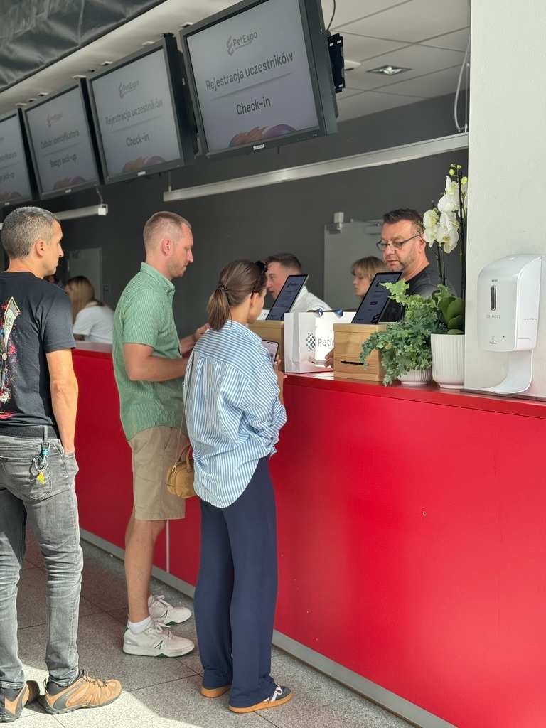 Ludzie stojący przy czerwonym stanowisku rejestracji na wydarzenie PetExpo z wyświetlaczami pokazującymi "Rejestracja uczestników Check-in".