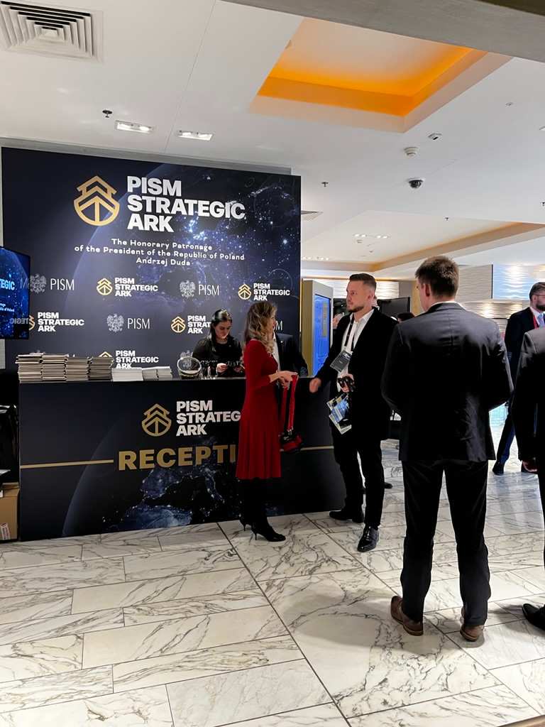 Stanowisko recepcyjne PISM Strategic ARK z osobami rozmawiającymi na marmurowej podłodze.