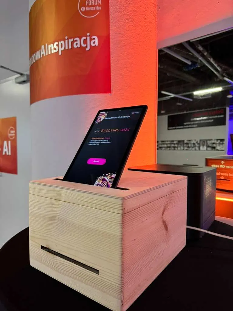 Tablet w drewnianej podstawce na tle pomarańczowej ściany z napisem Forum Horeca Idea.