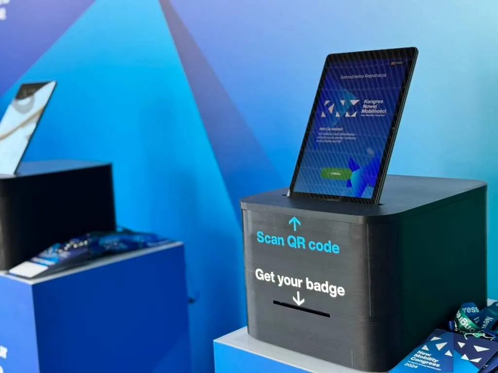 Stacja rejestracyjna z tabletem wyświetlającym Kongres Nowej Mobilności oraz napisem 'Zeskanuj kod QR, odbierz identyfikator'.