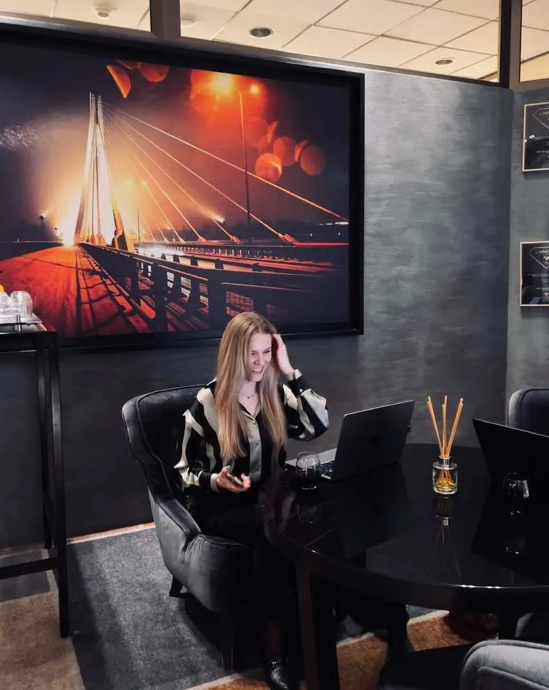 Kobieta siedząca przy czarnym stole z laptopem, obok kieliszek z czerwonym winem, na tle dużego zdjęcia mostu nocą.