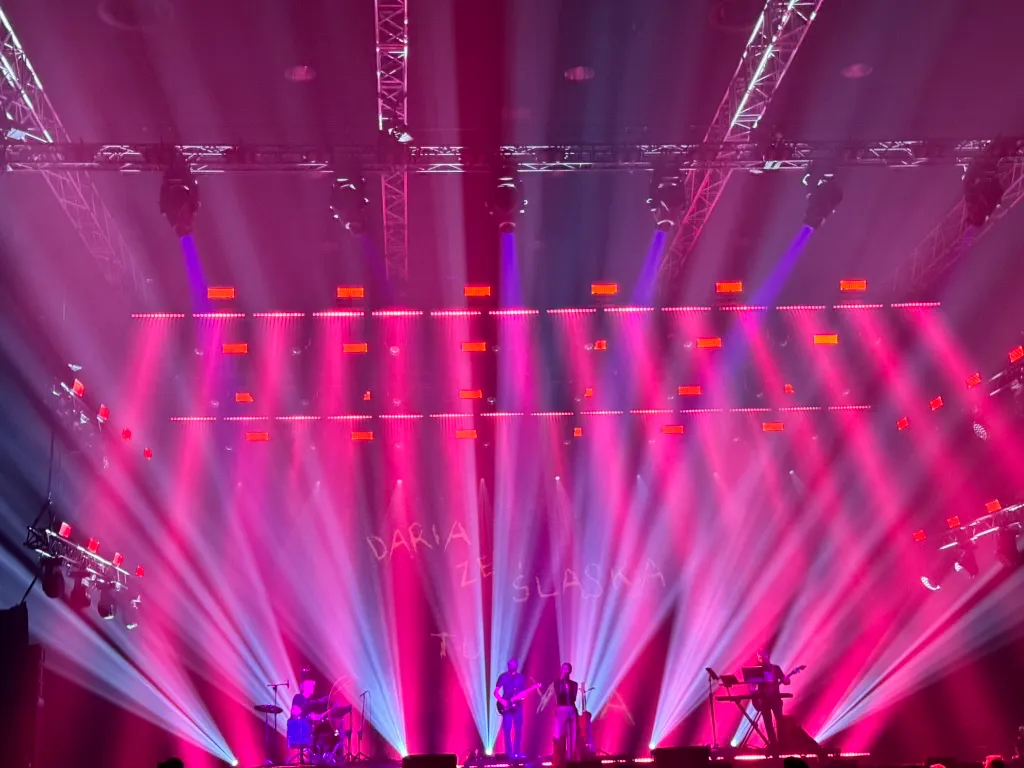 Zespół muzyczny na scenie z jasnym różowym i niebieskim oświetleniem.