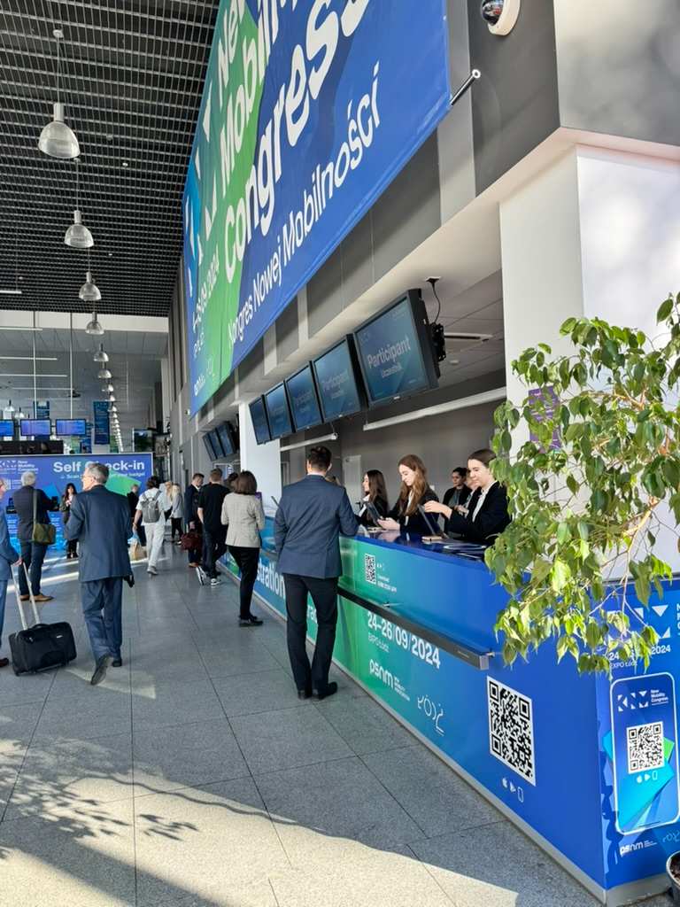 Ludzie rejestrujący się i rozmawiający przy stanowiskach recepcyjnych na konferencji lub targach w jasnym holu.