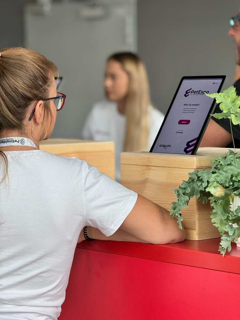 Kobieta w białej koszulce i okularach stoi przy czerwonym stole, rozmawiając z osobą po drugiej stronie, na stole stoi tablet z napisem PetExpo.