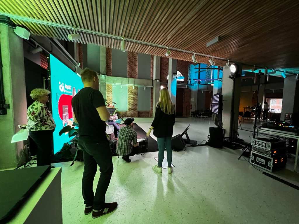 Grupa ludzi stoi i rozmawia w nowoczesnym wnętrzu z dużym ekranem LED i oświetleniem studyjnym.