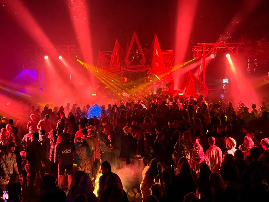 Tłum ludzi na nocnym koncercie z czerwonymi i żółtymi światłami sceny i niebieską kulą świetlną.