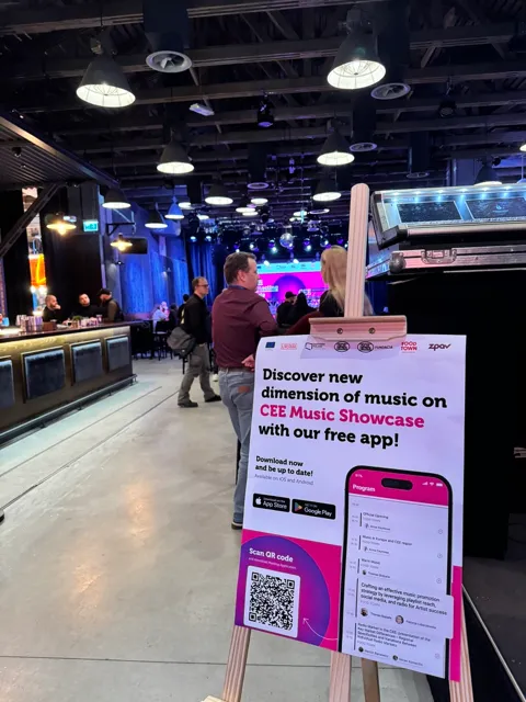 Plakat promujący bezpłatną aplikację CEE Music Showcase z programem wydarzenia, ustawiony na sztaludze w sali z oświetleniem i ludźmi w tle.