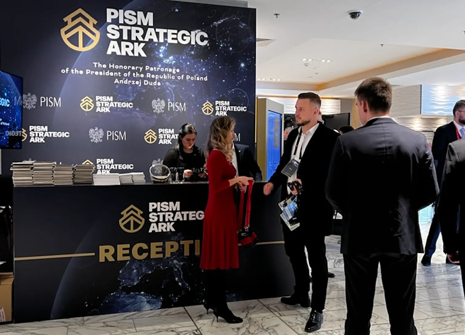 Recepcja PISM Strategic Ark z kilkoma osobami rozmawiającymi przed ladą rejestracyjną w eleganckim wnętrzu.