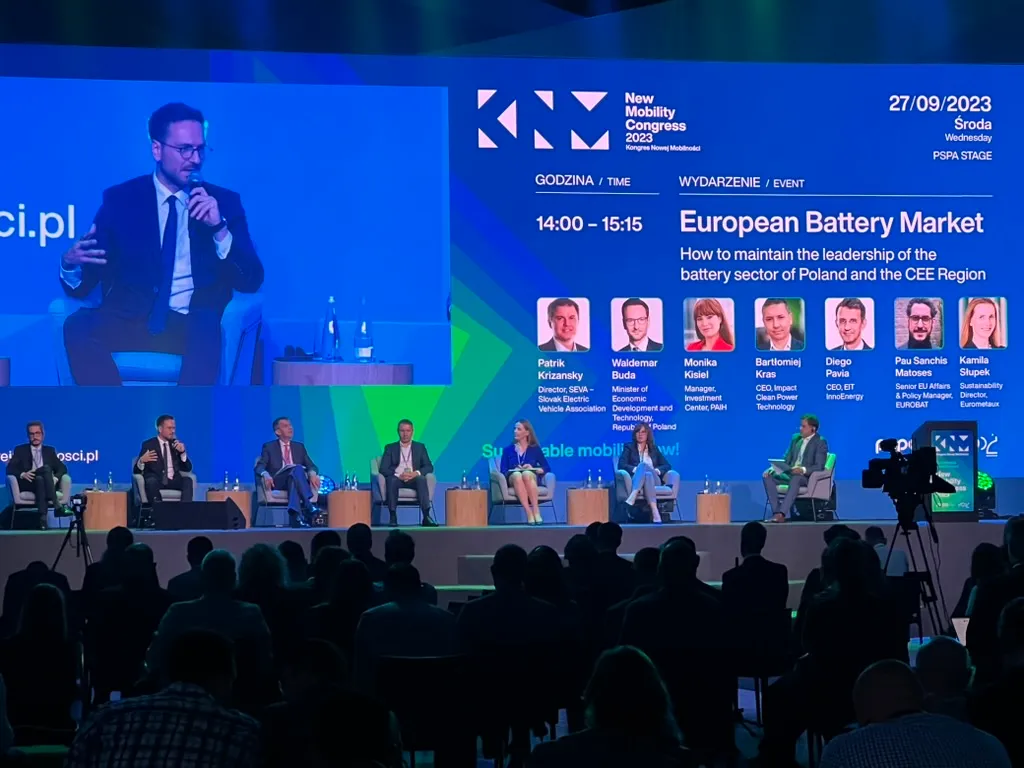 Panel dyskusyjny podczas New Mobility Congress 2023 o europejskim rynku baterii, z siedmioma prelegentami na scenie i dużym ekranem prezentującym szczegóły wydarzenia.