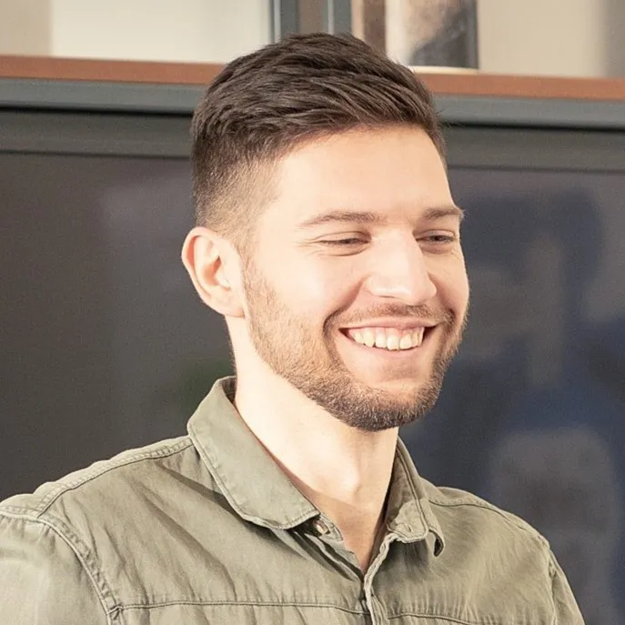 Mężczyzna z krótkimi ciemnymi włosami i zarostem, uśmiechający się, ubrany w zieloną koszulę.