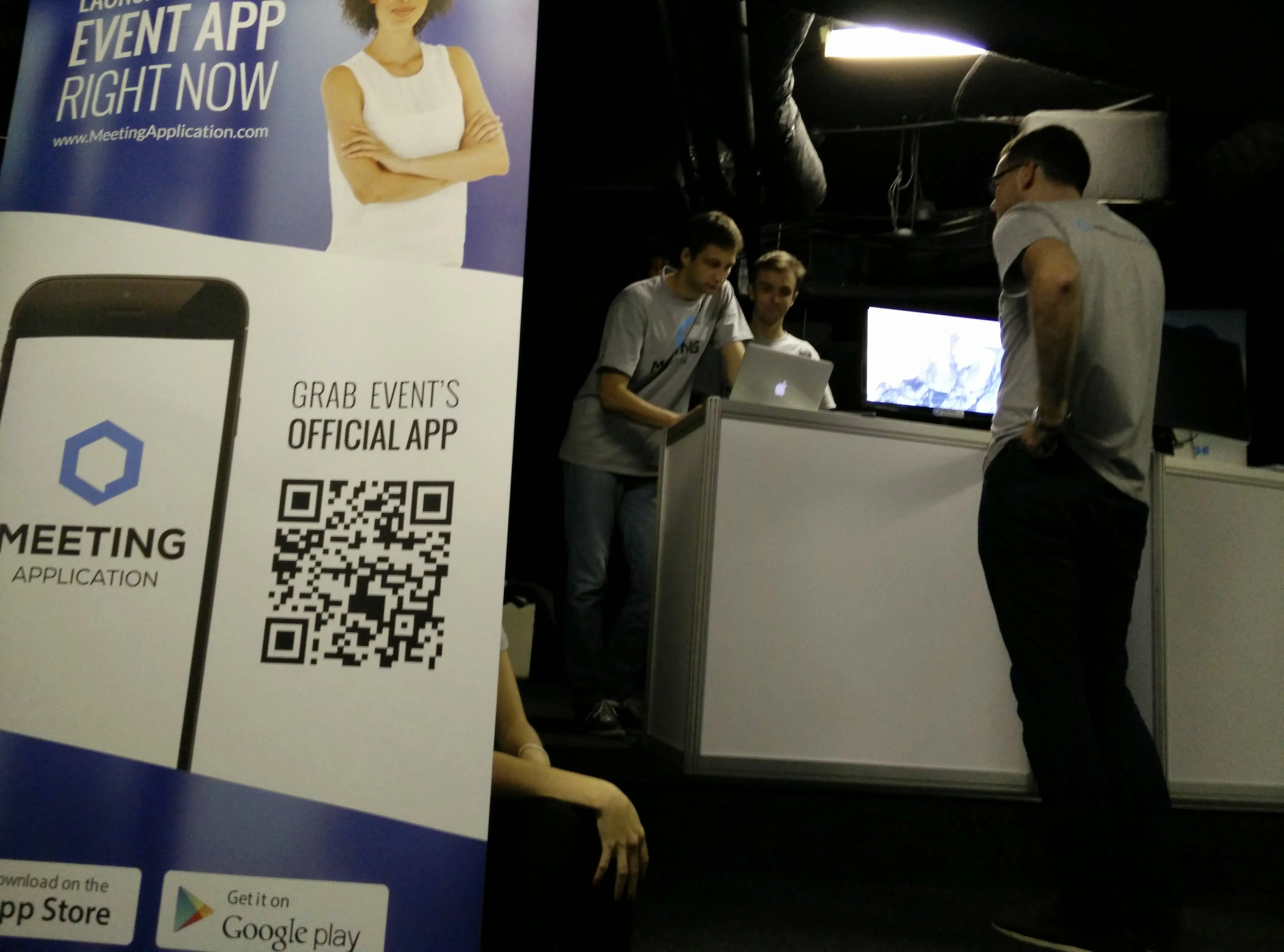 Stoisko promocyjne aplikacji Meeting Application z dużym banerem i trzema mężczyznami stojącymi przy stoisku z laptopem.