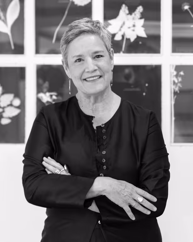 Smiling woman with short hair and earrings stands with arms crossed in front of floral artworks.