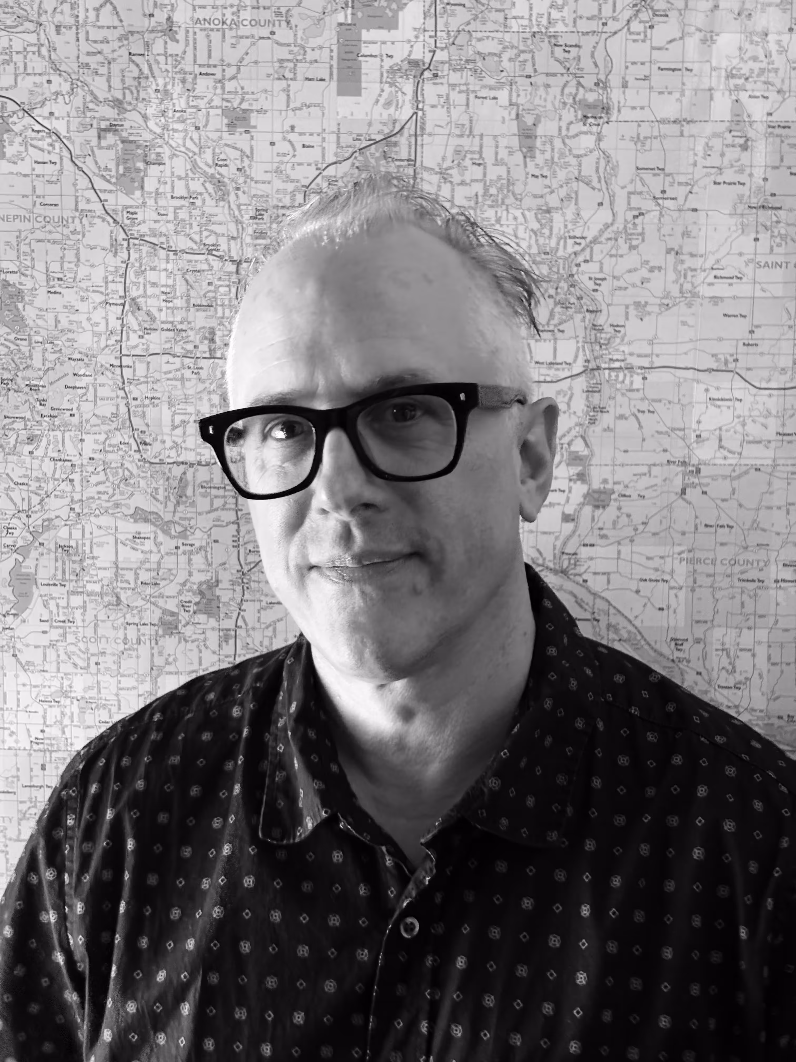 Man with glasses and patterned shirt in front of a detailed map background.