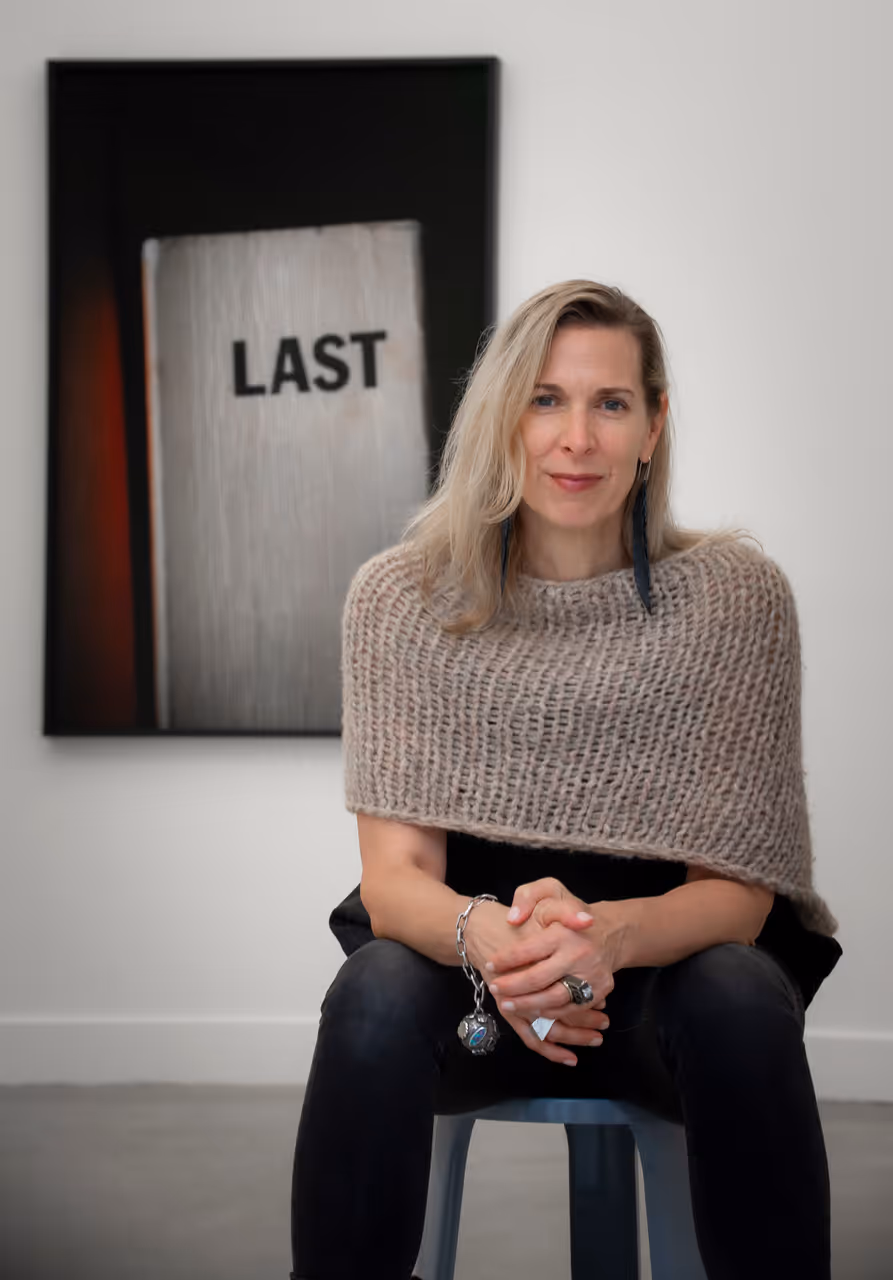 Blonde woman in a beige knitted shawl sits on a stool, with a framed 'LAST' artwork behind her.