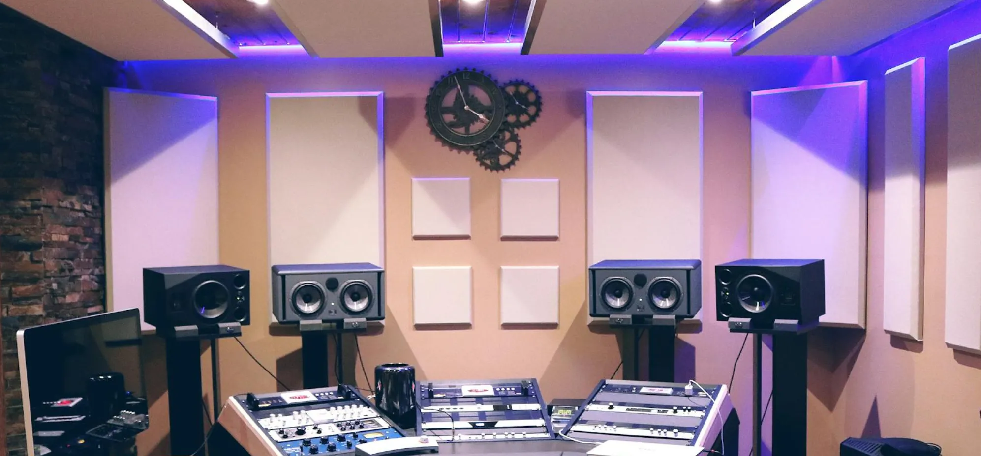 Professional audio mixing studio with speakers, control panels, acoustic panels, and a gear-themed wall clock under purple ambient lighting.