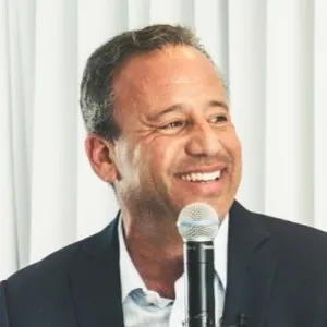Smiling man in a dark suit speaking into a microphone with white curtains in the background.