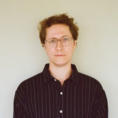Man with light skin and curly brown hair wearing glasses and a black striped button-up shirt against a plain light background.