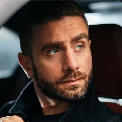 Bearded man with dark hair looking thoughtfully to the side inside a car.