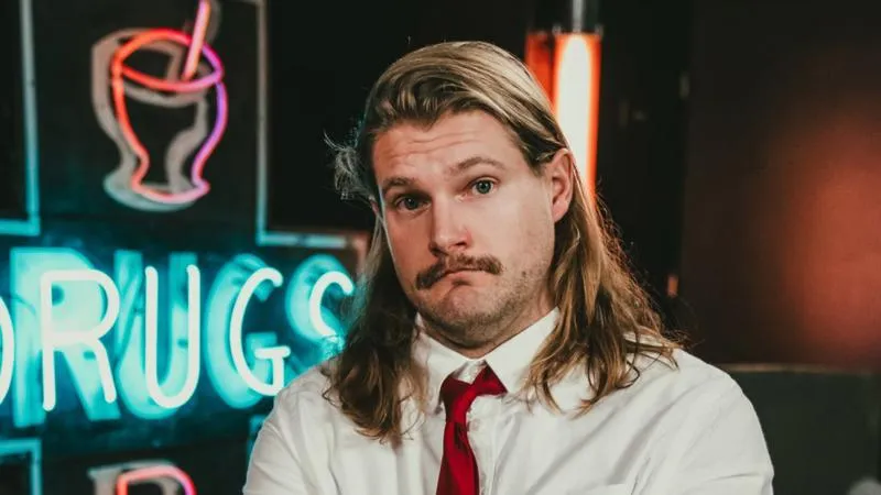 Man with long blonde hair and a mustache wearing a white shirt and red tie in front of a neon sign with the word 'DRUGS'.