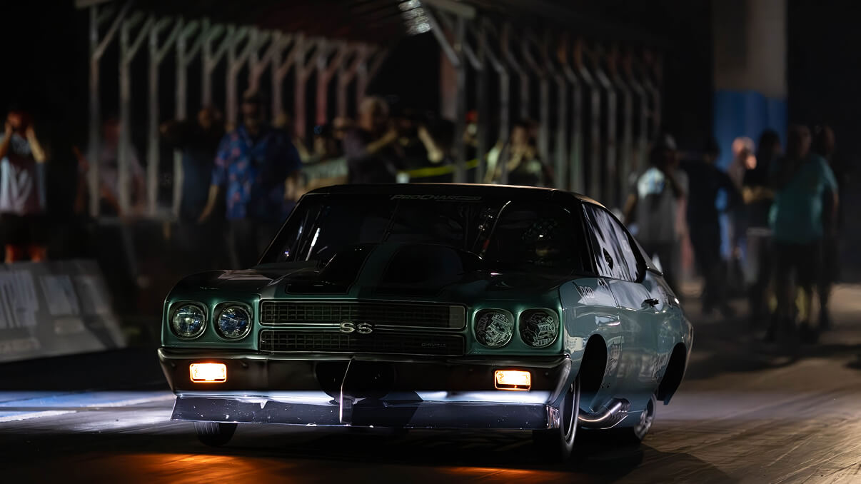 Green Chevrolet SS muscle car racing on a drag strip at night with spectators behind a fence.