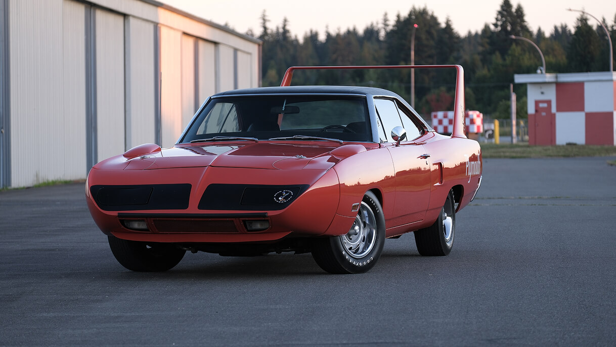 Red 1970 Plymouth Superbird parked on pavement near a building and trees.